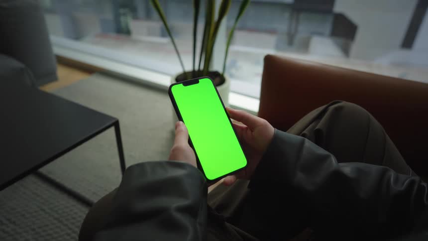 A person holds a smartphone with a green screen while relaxing in a modern armchair indoors