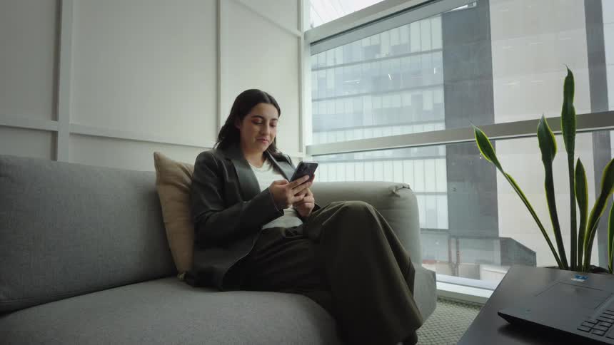 A young woman relaxes on a gray couch in a modern office lounge, smiling as she scrolls through her smartphone