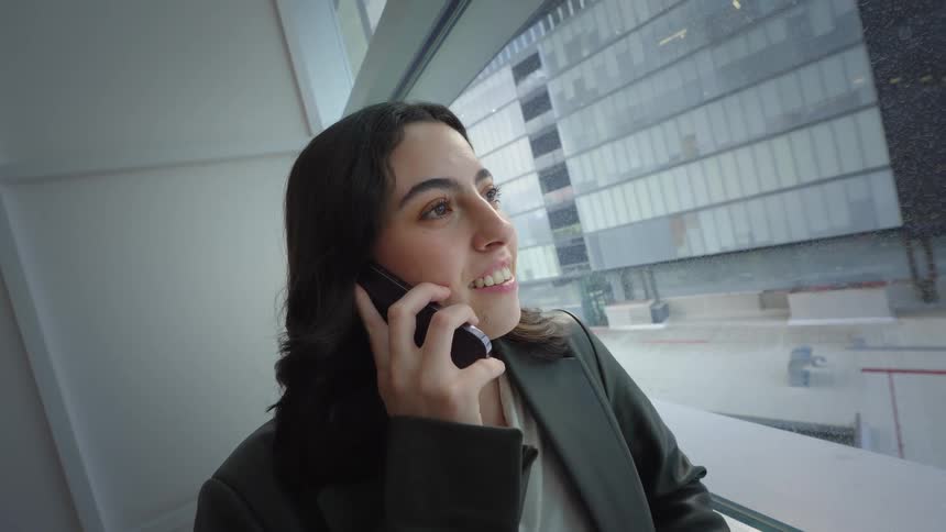 A young businesswoman smiles while making a phone call on her smartphone next to a large window overlooking the city