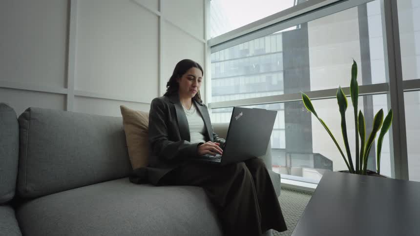 A smiling professional woman works on her laptop while sitting on a comfortable grey couch in a modern office lounge