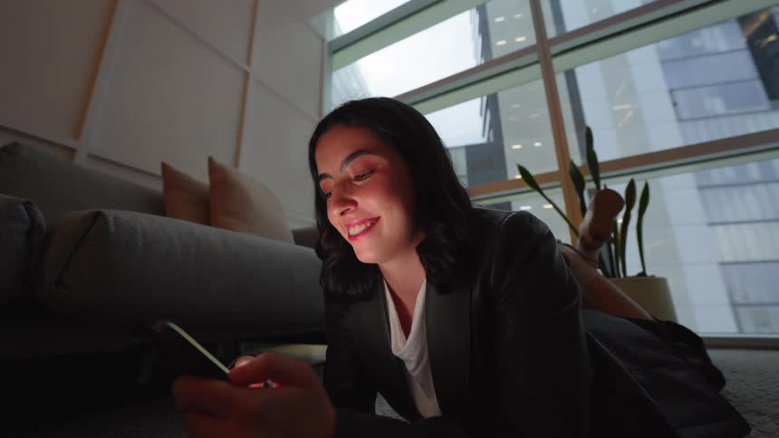 A happy businesswoman in a suit relaxes on the floor of her modern high-rise apartment at night, smiling while using her smartphone