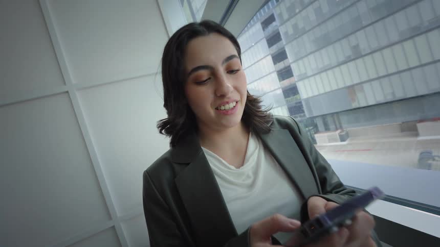 A smiling young professional woman texts on her smartphone while standing by a large window in a modern city office