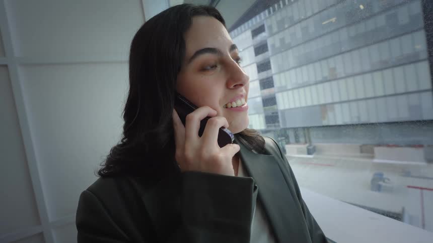 A young professional woman smiles while talking on her cell phone, standing by a large window with a city view