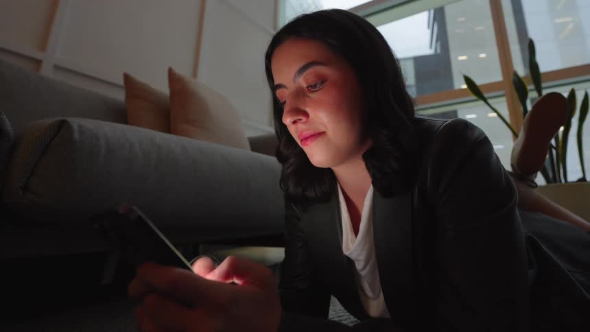 A young businesswoman relaxes on the floor of a modern office at night, smiling as she scrolls through her smartphone