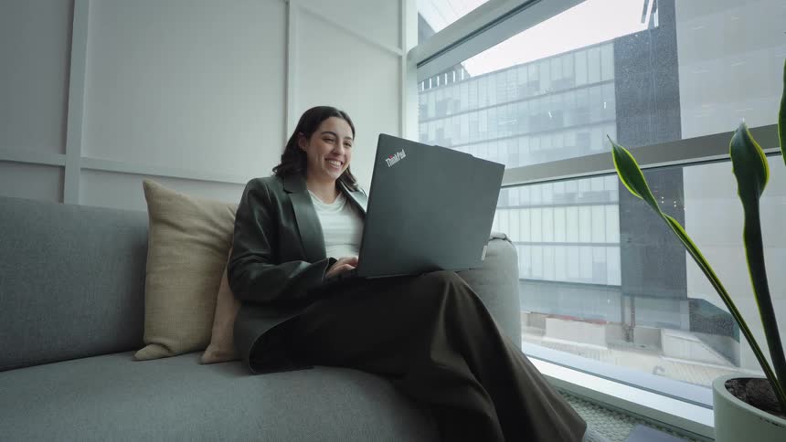 A happy young businesswoman smiles while working on her laptop from a comfortable couch in a modern office