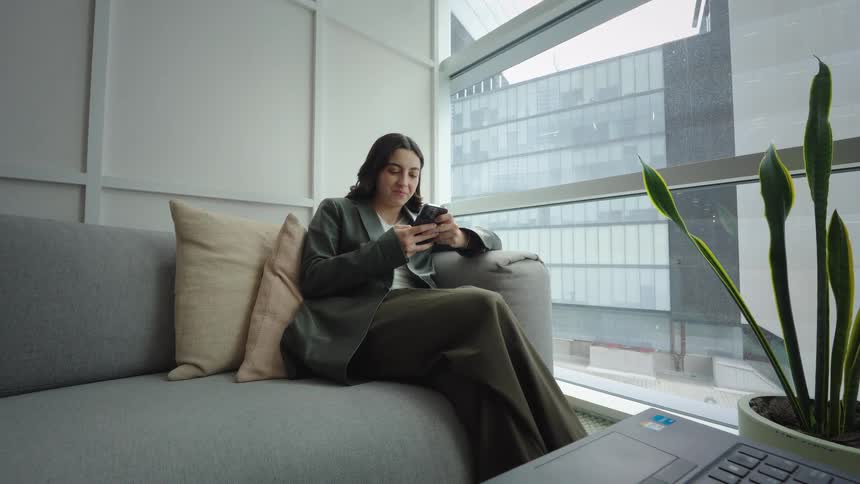 A young businesswoman relaxes on a sofa in a modern office lounge, smiling as she uses her smartphone