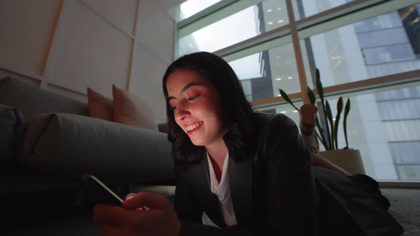 A happy businesswoman in a suit relaxes on the floor of a modern city high-rise at night, smiling as she looks at her smartphone