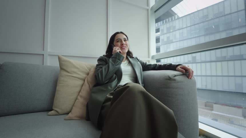 A young woman relaxes on a comfortable couch in a modern office lounge, smiling as she talks on her smartphone