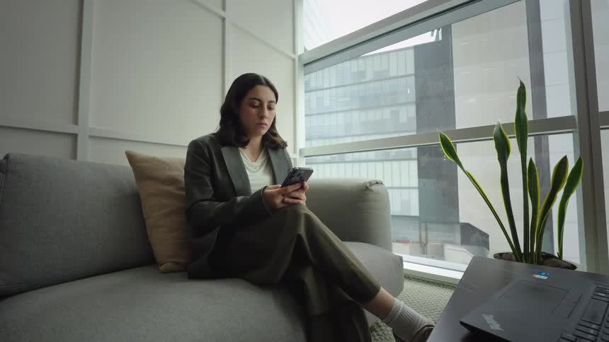 A professional woman in a modern high-rise office relaxes on a sofa, scrolling on her phone before answering a call