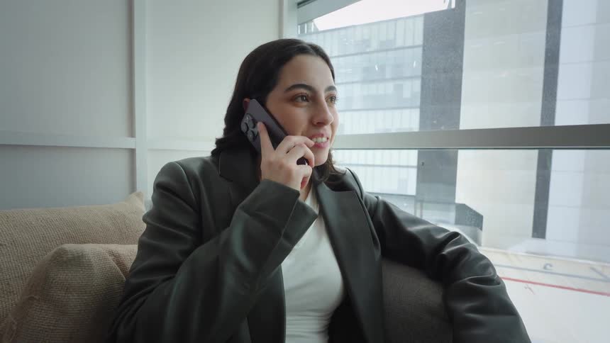 A young businesswoman smiles while answering a phone call as she relaxes on a couch in a modern high-rise office