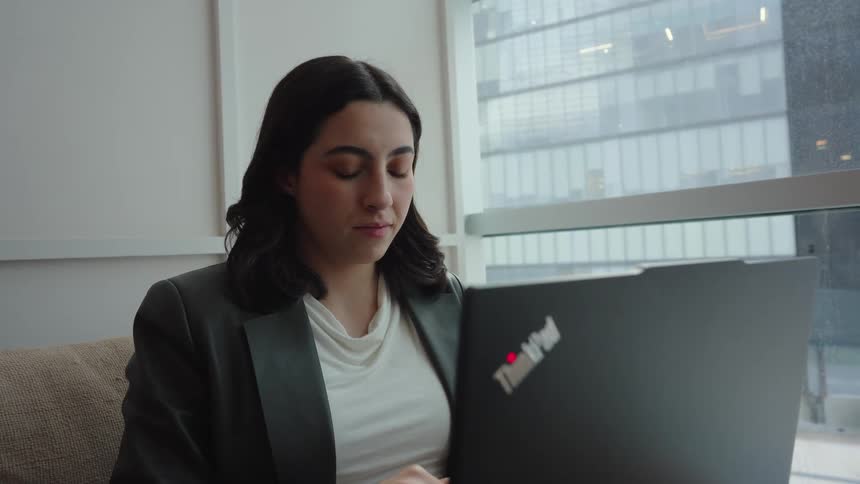 A focused businesswoman works on her laptop while sitting on a couch in a modern office with a city view