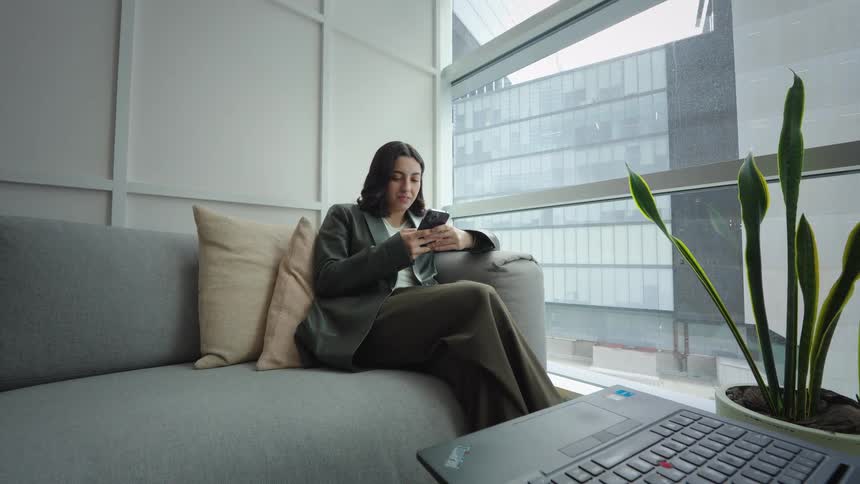A young professional woman smiles while texting on her phone as she relaxes on a couch in a modern office lounge