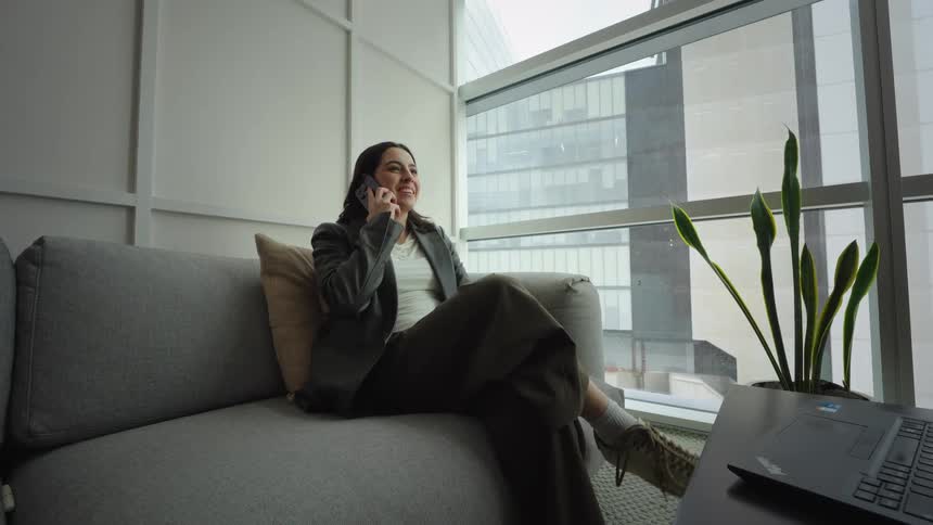 A young businesswoman smiles while taking a phone call, relaxing on a couch in a modern office with a city view