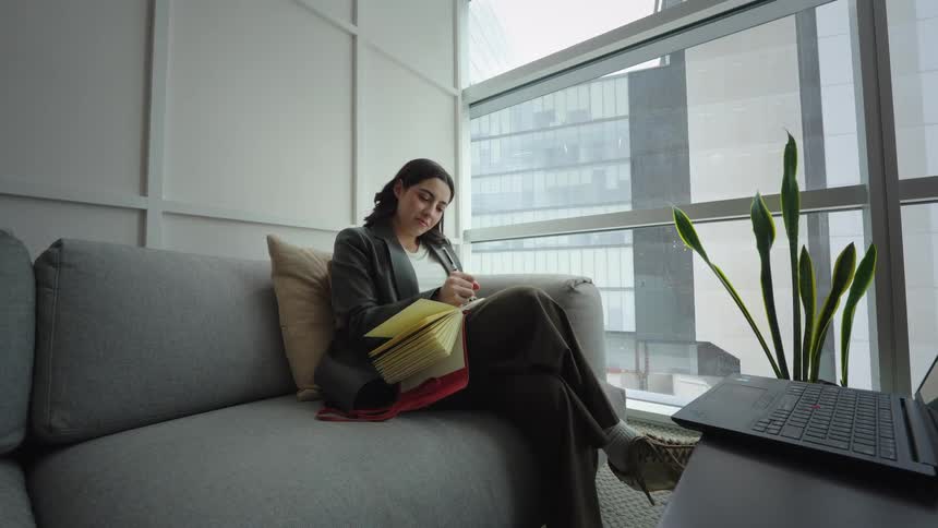 A businesswoman sits on a couch in a modern office with a city view, writing in her journal