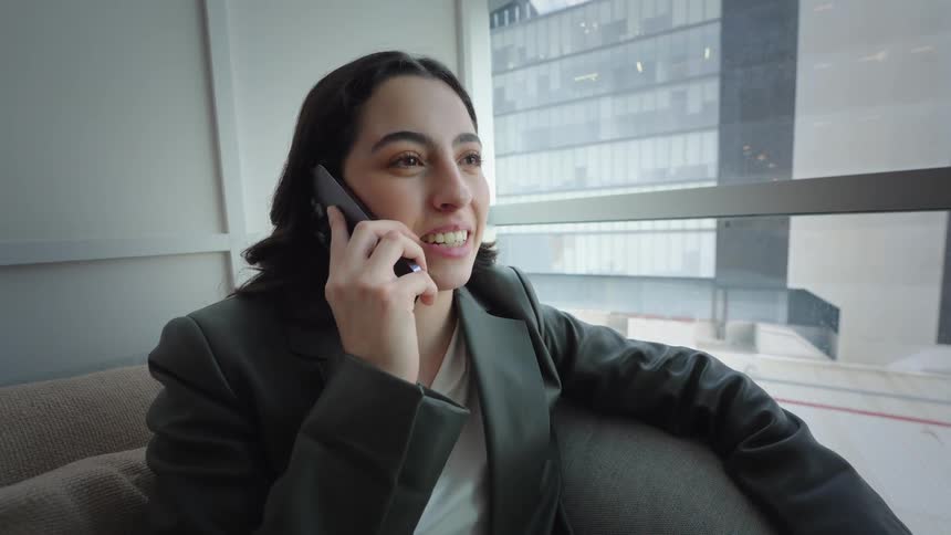 A smiling businesswoman talks on her smartphone while relaxing on a couch by a large window in a modern city office