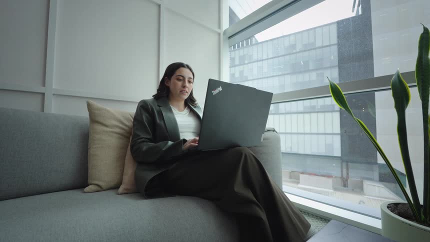 A young professional woman works on her laptop while comfortably sitting on a couch in a modern office with a city view