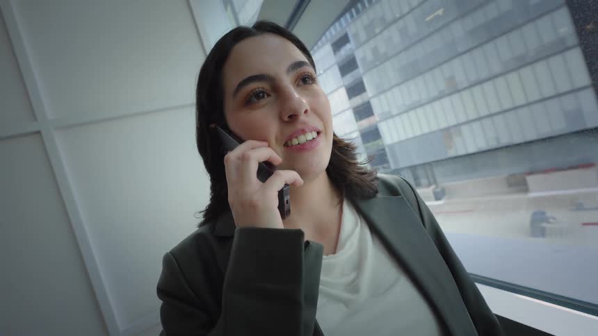 A young businesswoman smiles while taking a phone call next to a large window in a modern office as the camera moves in