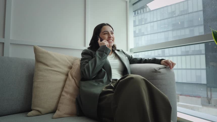 A young businesswoman smiles while having a pleasant phone conversation on a sofa in a modern city office