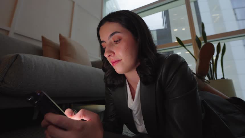 A businesswoman lies on the floor in a modern room and scrolls on her smartphone, its screen illuminating her face in the dim light