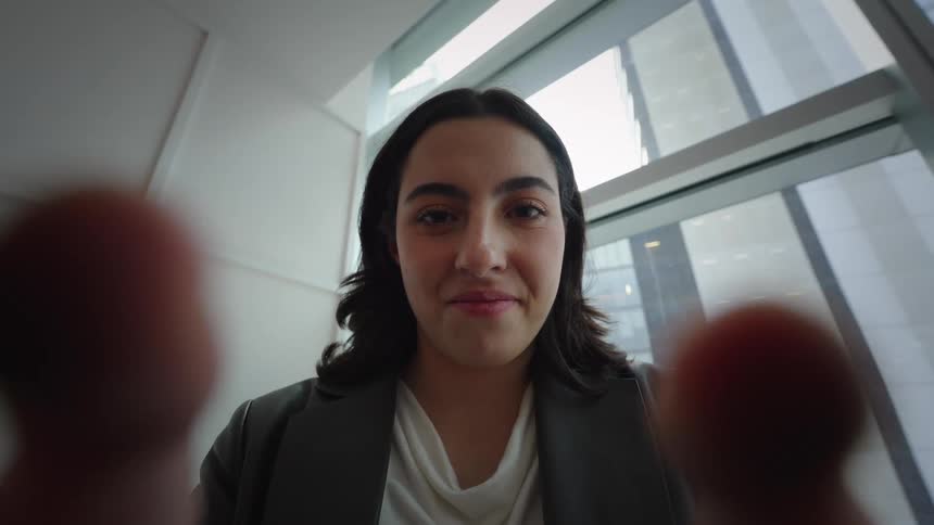 First-person view of a young businesswoman holding a phone and smiling at the camera in a modern high-rise office