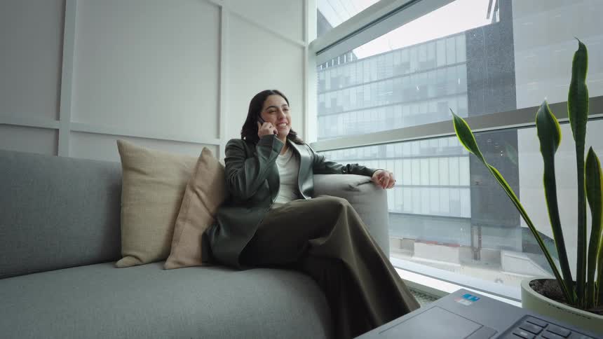 A happy businesswoman takes a pleasant phone call while relaxing on a couch in a modern office with a city view