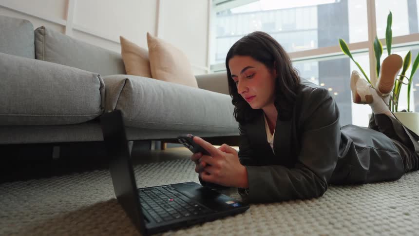 A young businesswoman lies comfortably on the floor of a modern city apartment, scrolling through her smartphone with her laptop nearby