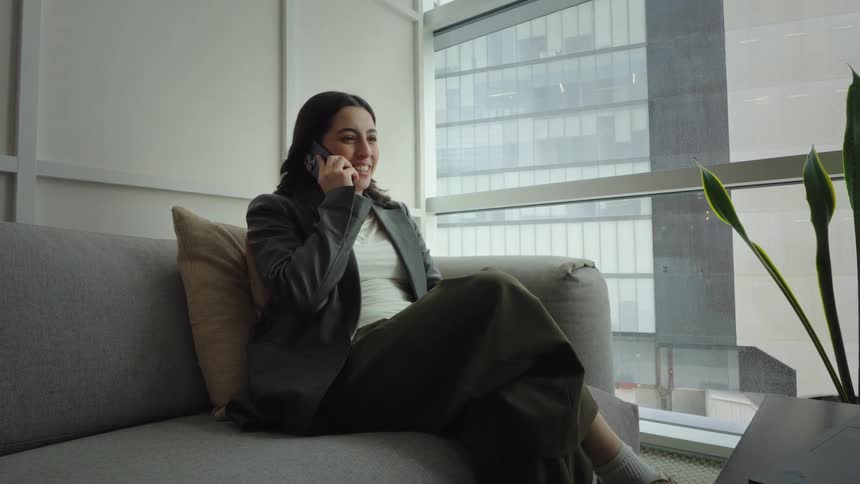 A relaxed young professional woman smiles while talking on her phone, comfortably seated on a couch in a modern high-rise office