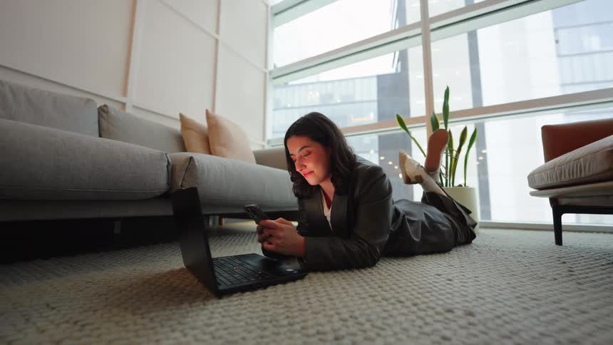A young businesswoman lies on the floor of her modern apartment, smiling as she scrolls through her smartphone with her laptop nearby
