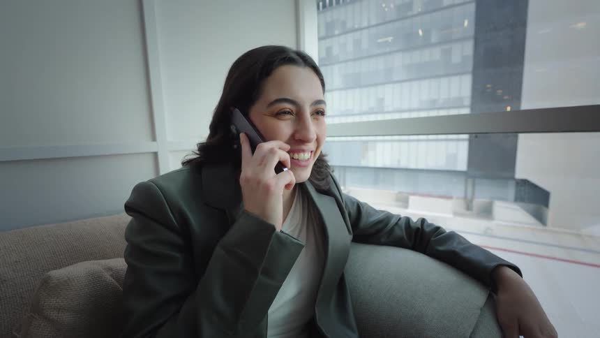 A young professional woman smiles happily while talking on her smartphone, seated on a comfortable couch in a modern office lounge