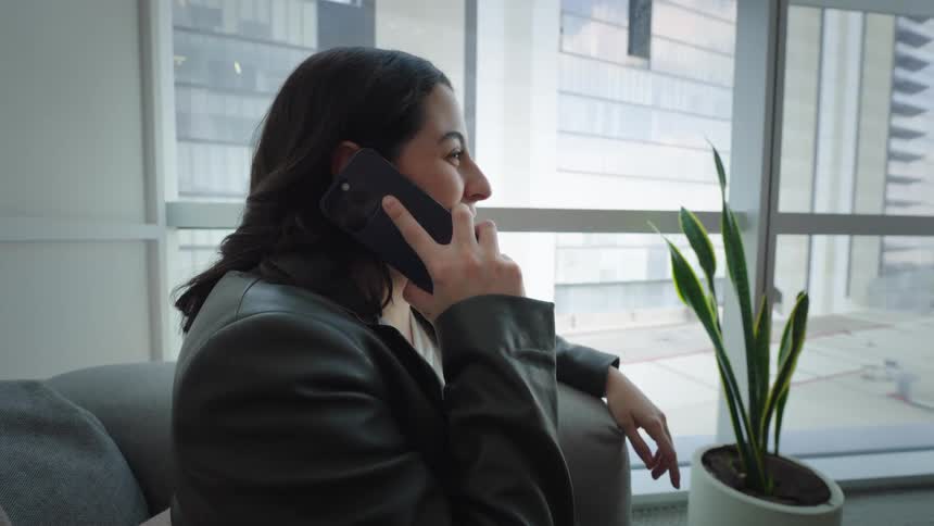 Young woman answers a phone call while relaxing on a couch in a modern high-rise apartment with city views