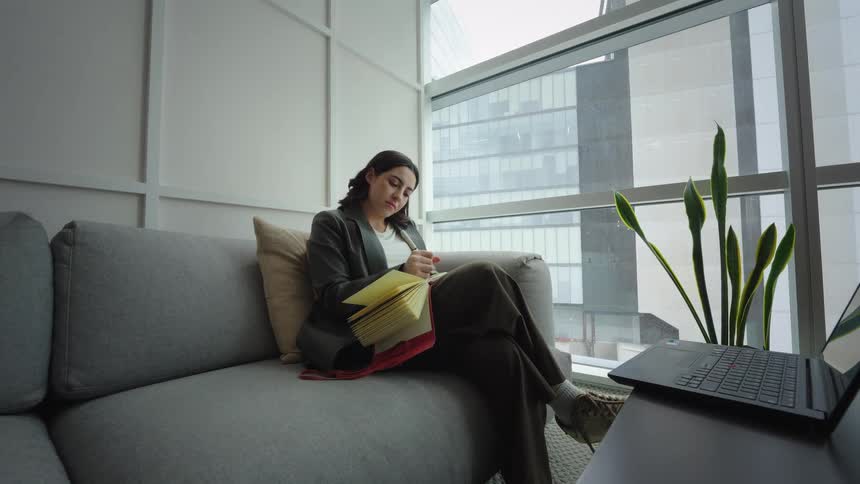 Slow pull-out shot of a young professional woman taking notes in a journal while sitting on a couch in a modern office with a large window view of the city