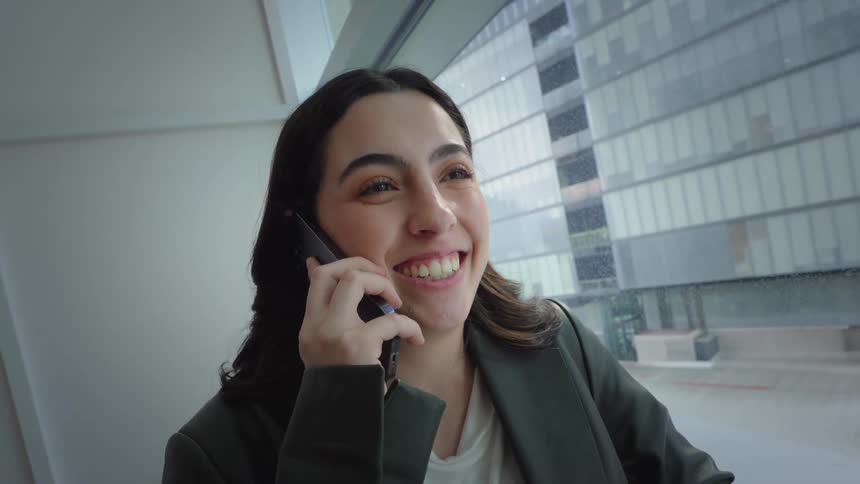 A smiling young businesswoman has a pleasant conversation on her smartphone next to a large office window