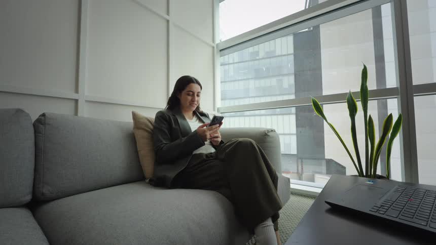 A smiling young woman sits comfortably on a sofa in a modern apartment, happily scrolling through her smartphone
