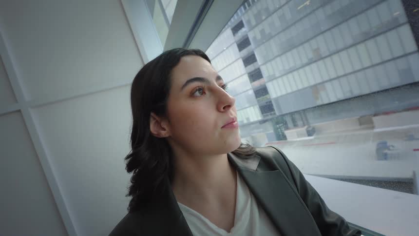 A professional woman looks up from her phone and smiles hopefully while looking out a large window in a modern city office