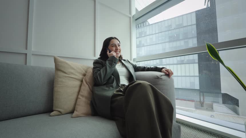 A young professional woman comfortably sits on a couch in a modern office with city views, happily talking on her smartphone