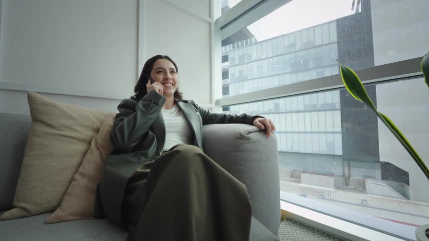 Low angle shot of a smiling businesswoman relaxing on a comfortable couch in a modern high-rise office while on a phone call