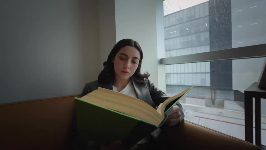A focused young woman in a blazer reads a large book on a leather sofa next to a city view window