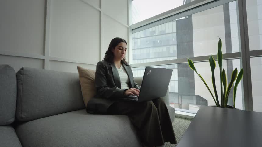 A focused businesswoman works on her laptop while sitting on a comfortable couch in a modern office lounge