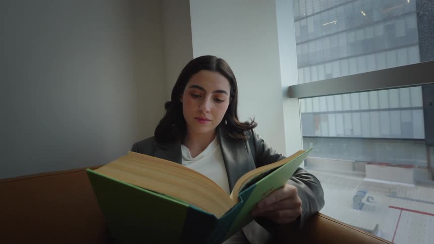 A focused young woman in a blazer reads a hardcover book while sitting on a brown sofa next to a large window overlooking the city