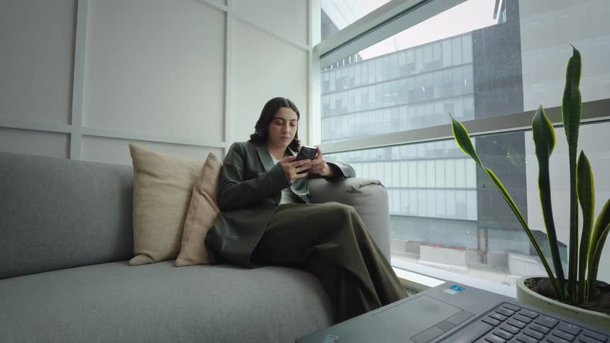 A professional woman smiles while scrolling on her smartphone, relaxing on a couch in a modern office with a city view