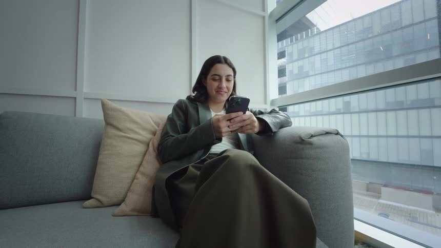 A stylish young woman sits on a modern couch by a large window in a city building, smiling as she texts on her smartphone