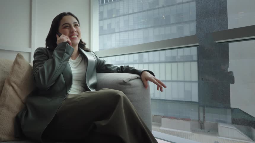 A stylish young woman smiles and talks on her cell phone while relaxing on a couch next to a window with a city view