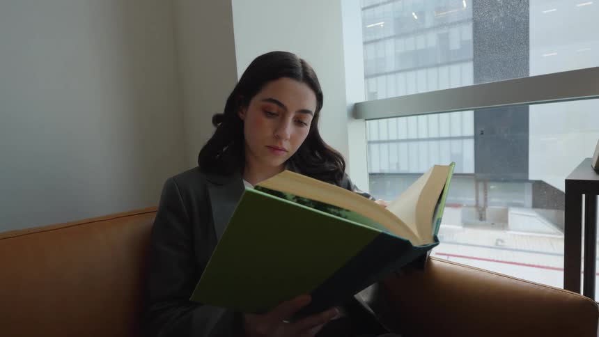 A focused young woman in a blazer sits on a leather couch and reads a large book next to a window overlooking the city