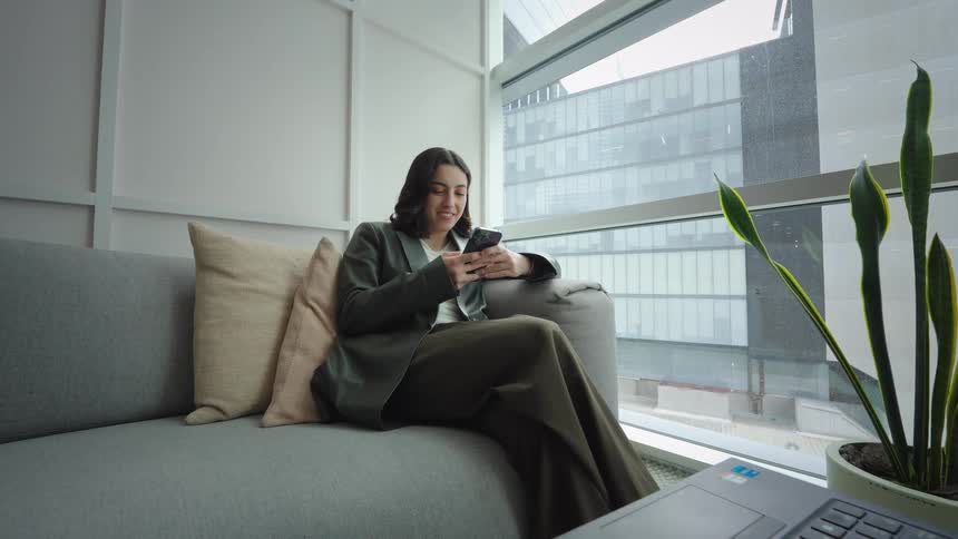 A young professional woman smiles while using her smartphone on a comfortable sofa in a modern office with a large window view of the city