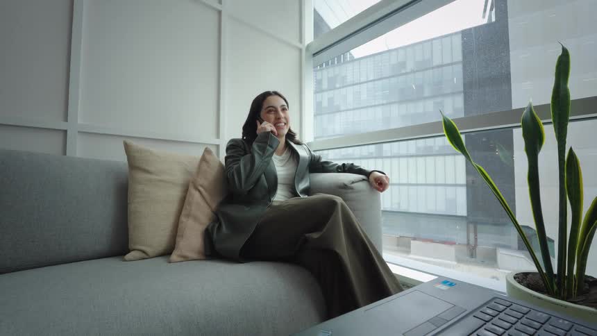 Smiling businesswoman relaxes on a couch in a modern high-rise office while talking on her smartphone as the camera pulls back