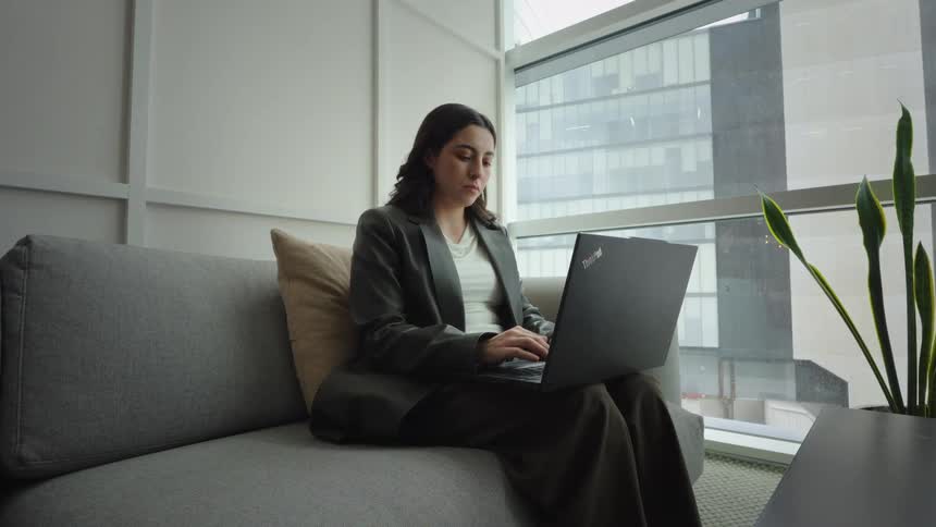 A young businesswoman works diligently on her laptop from a couch in a contemporary high-rise office with a city view