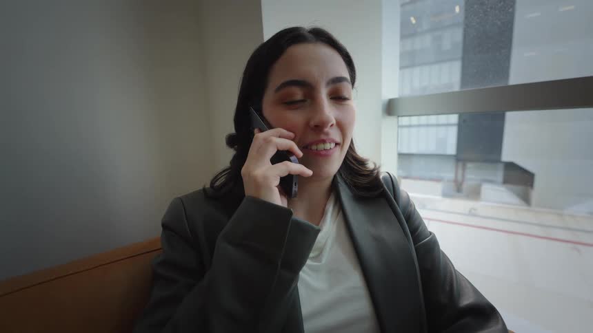 A young professional woman smiles while answering a phone call on a couch in a modern city office lounge