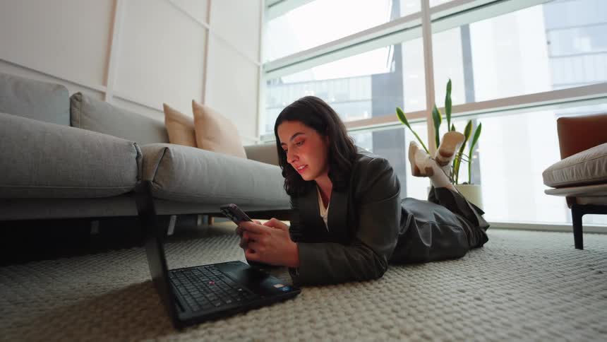 A young businesswoman smiles while using her smartphone, lying on the floor next to her laptop in a modern, sunlit living room