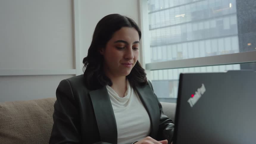 A smiling young professional woman works on her laptop while sitting comfortably on a couch in a modern office