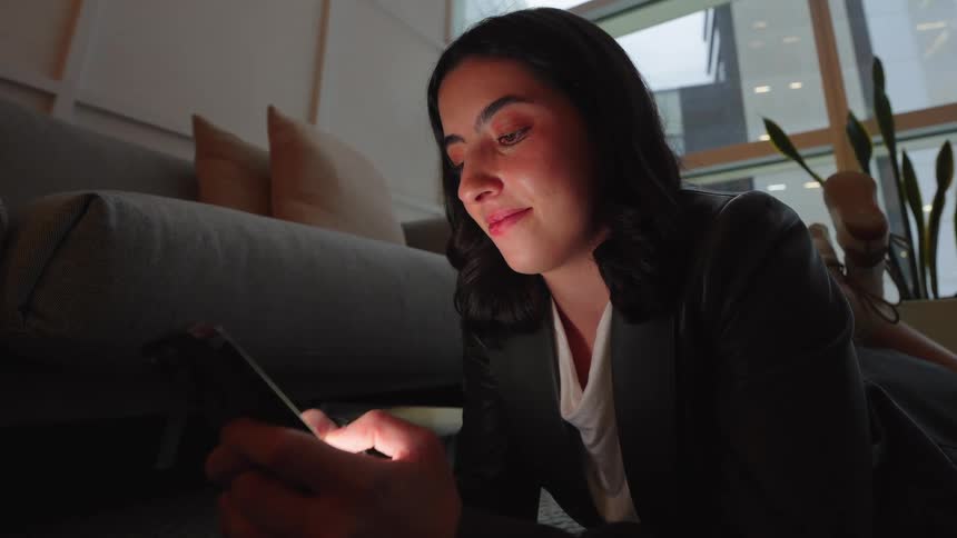 A young woman relaxes on the floor in a dimly lit room, her face illuminated by the glow of her smartphone as she smiles